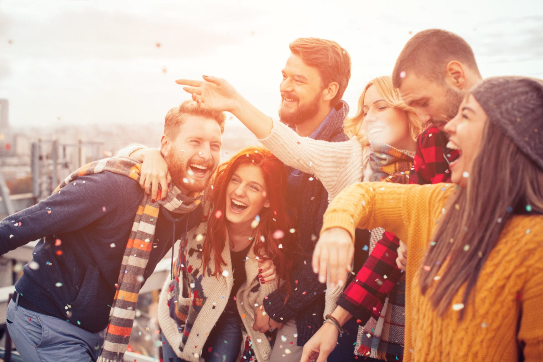 Couple of friends having a party on the rooftop of the building. Dancing and trowing a confetti in the air. Wearing knitted sweaters, hats and scarfs. They are happy and joyful.