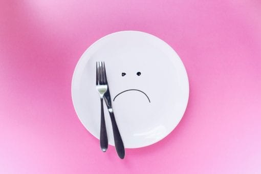 Photo of a white plate with a ad face drawn on it, with a knife and fork resting on top, in front of a pink background