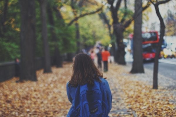 A child with long brown hair, blue backpack and blue jacket walking away from the camera on a sidewalk with fallen leaves.