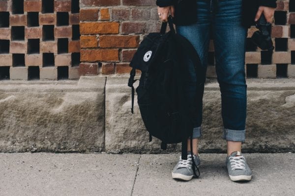 Photo of a person's legs, wearing jeans and sneakers, holding a backpack