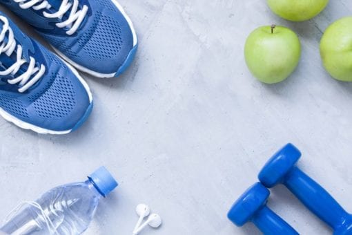 Blue sneakers, hand weights, apples and a water bottle laying flat on a gray background