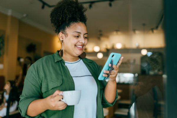 Happy curly haired women in cafe holding coffee and looking at her phone